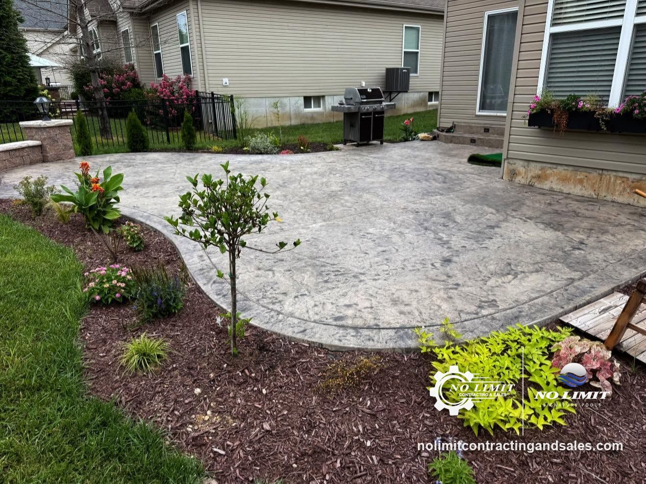 Gray stamped concrete patio curving around landscaped beds by No Limit Contracting & Sales