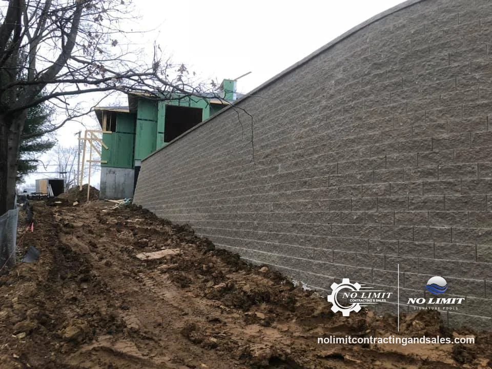 Tall gray block retaining wall beside house under construction, muddy backfill by No Limit Contracting & Sales