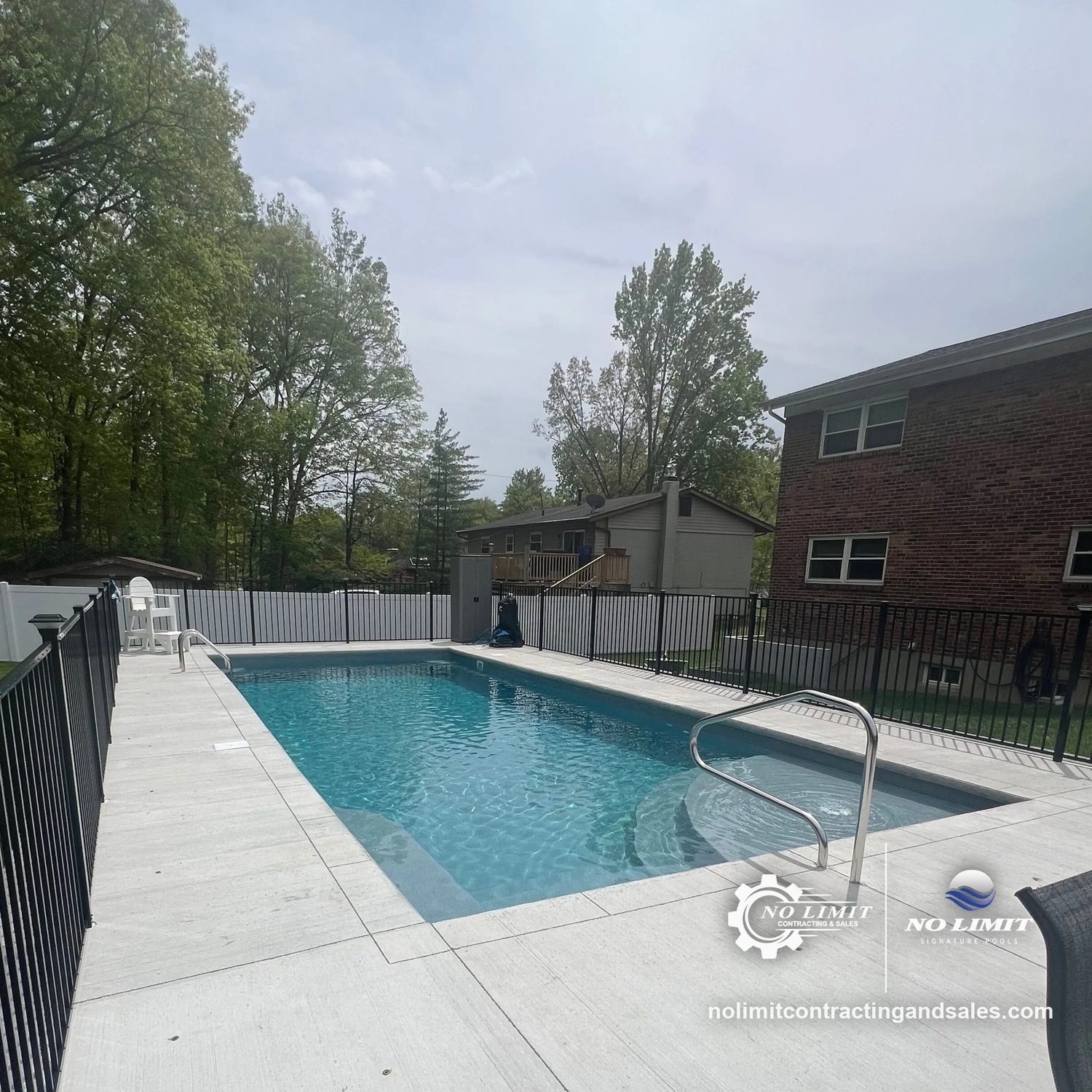 Finished rectangular fiberglass pool with a light deck and black fence beside a brick house by No Limit Signature Pools