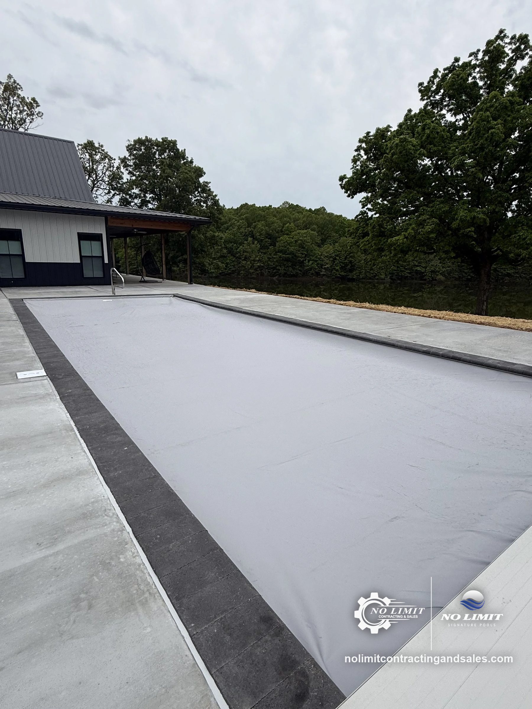 Rectangular pool under gray automatic/safety cover, concrete deck, lake behind by No Limit Signature Pools